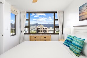 A bedroom with a large window, a bed, a wooden dresser, and a view of a cityscape. - Waikiki Banyan condo w/ pool & beach access (Honolulu)