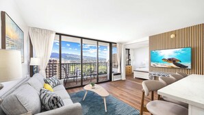 A modern living room with a large TV, a sofa, a dining area, and a balcony with a city view. - Waikiki Banyan condo w/ pool & beach access (Honolulu)