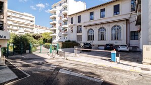 Un edificio residencial de varios pisos con entrada porticada, un área de estacionamiento con automóviles y un cielo despejado.