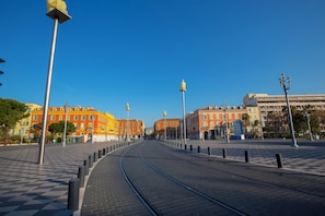 Une voie ferrée de tramway mène à une place dont le sol est pavé de motifs en damier et où s’alignent des bâtiments à la façade jaune.