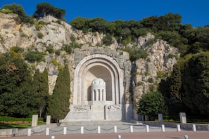 Un monument de pierre orné d’inscriptions, entouré d’un petit muret et d’une chaîne de protection, situé au pied d’une colline rocheuse et bordé d’arbres.