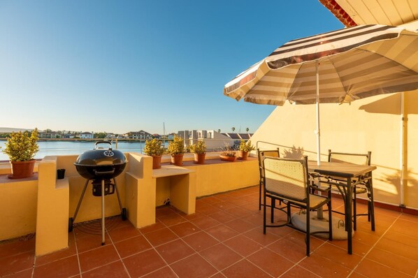 A terrace with a grill, potted plants, a table with chairs, and a striped umbrella. - Luxury Riverside Views | Balcony | Pool (Velddrif)