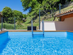 A swimming pool with a wooden deck, metal railings, and a stone wall in the background.