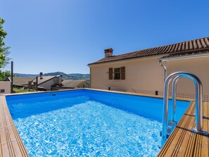 A swimming pool with a wooden deck, a metal ladder, and a view of buildings and hills in the background.