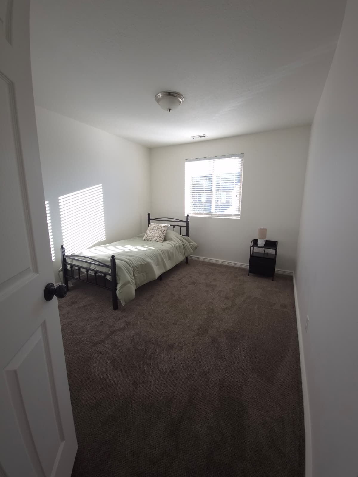 Une chambre avec un lit simple, une table de chevet, une fenêtre avec des stores et une porte.