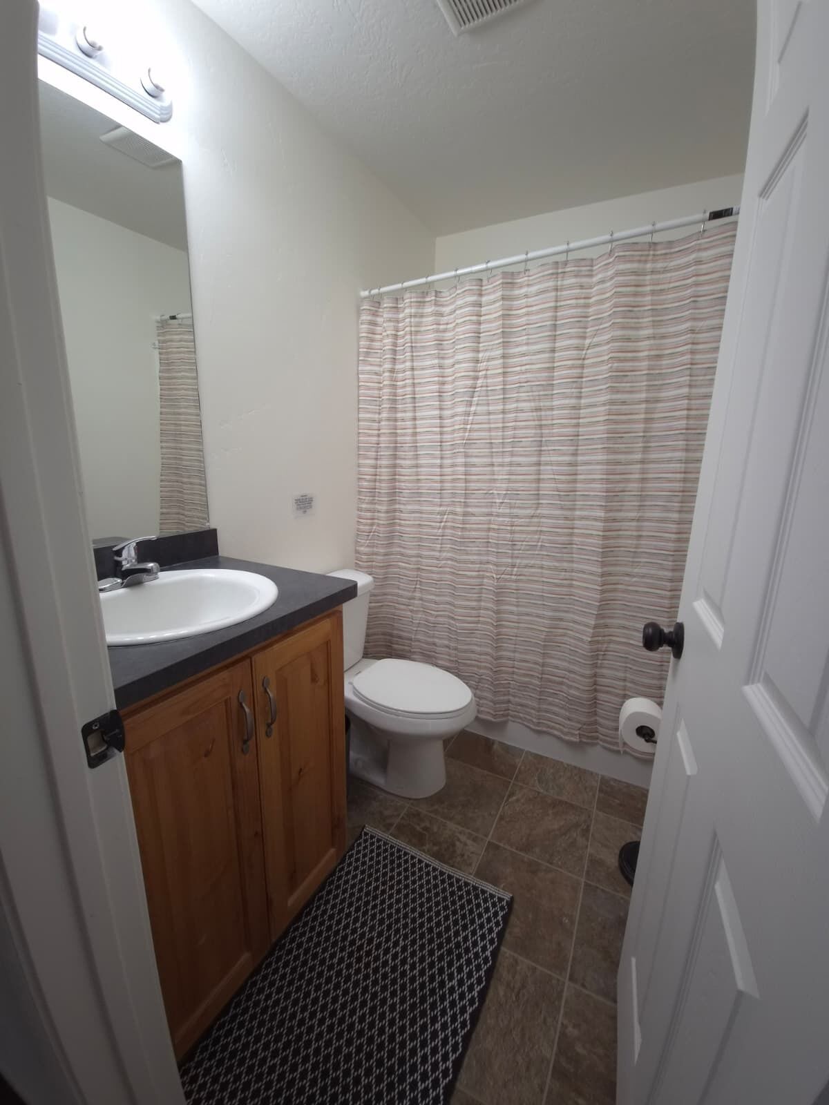 A bathroom with a white sink, wooden cabinets, a toilet, a striped shower curtain, and a houndstooth rug.