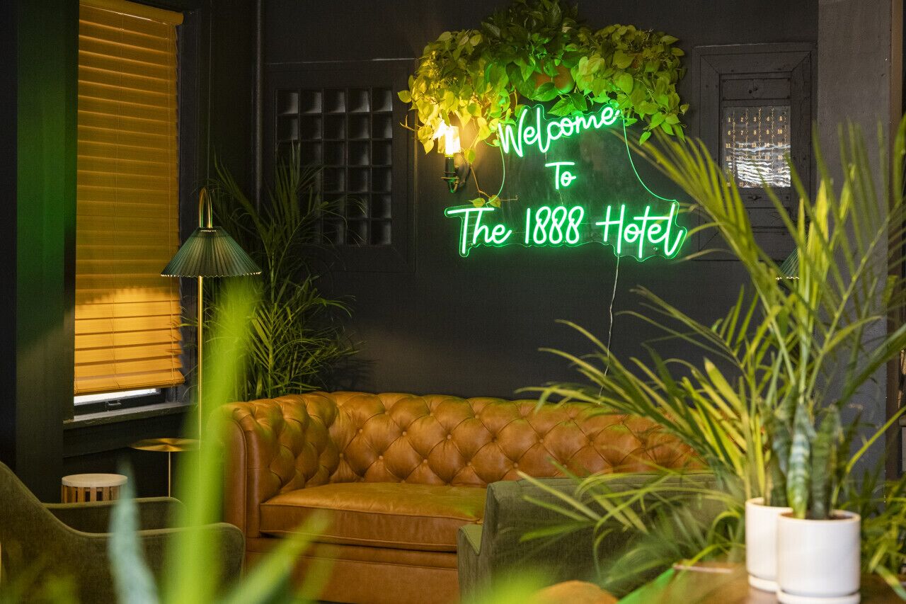 A neon sign reading "Welcome to The 1888 Hotel" above a leather sofa in a warmly lit room with green plants.