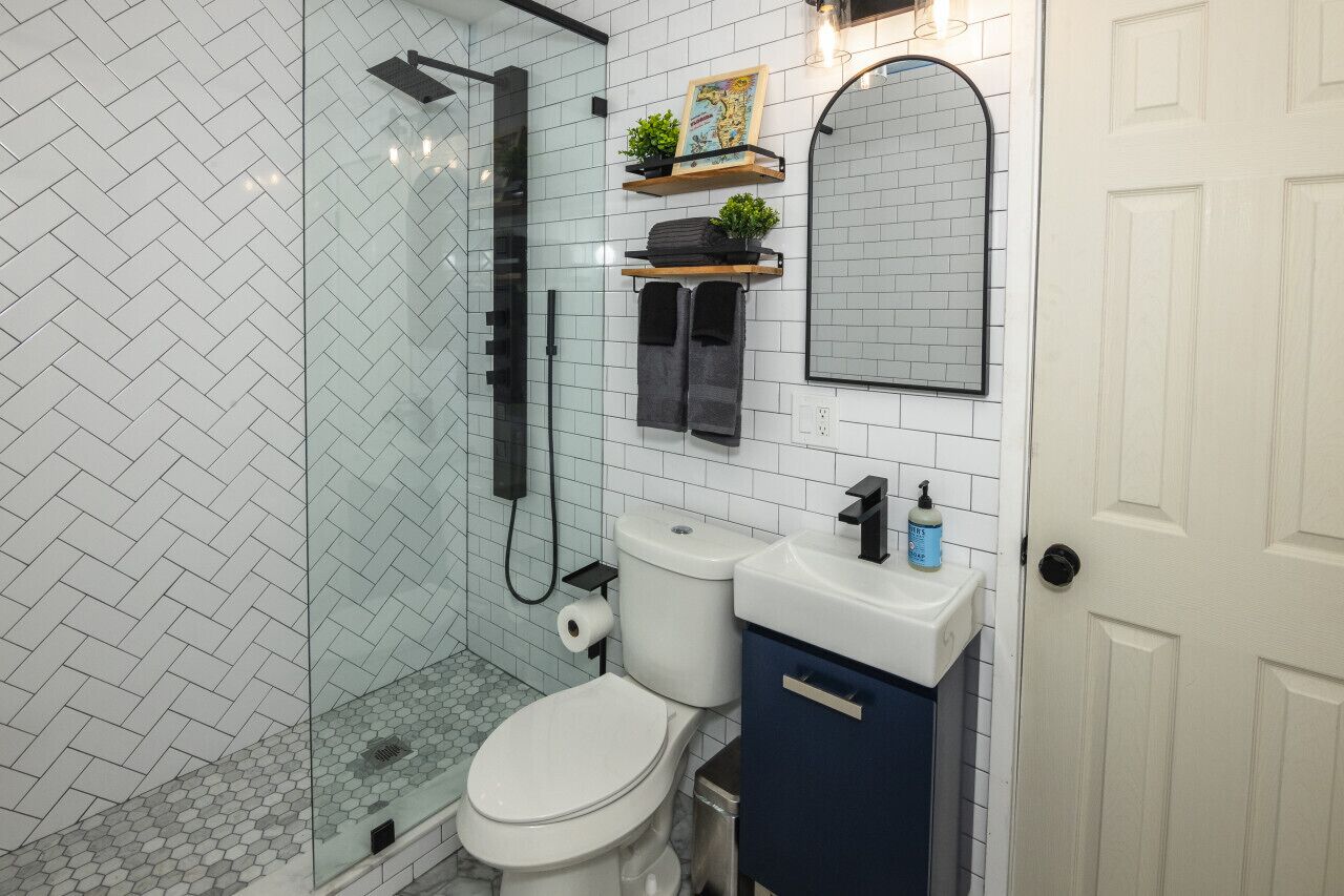 A bathroom with a glass shower enclosure, a white toilet, a sink with a blue soap dispenser, and a mirror above the sink.