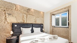 Una camera da letto con parete in pietra, un letto con biancheria bianca, due cuscini, un comodino con una lampada e una finestra con vista.