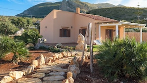 Ein mediterranes Haus mit einem Kiesweg, der zum Eingang führt, umgeben von viel Grün und einer Bergkulisse.