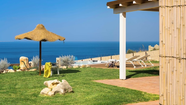 Ein Poolbereich mit einem Reetdachschirm, Liegestühlen und Blick aufs Meer.