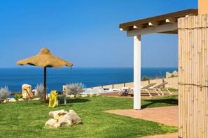 Un espace aménagé au bord de la piscine, avec un parasol en chaume, des chaises longues et une vue sur la mer.