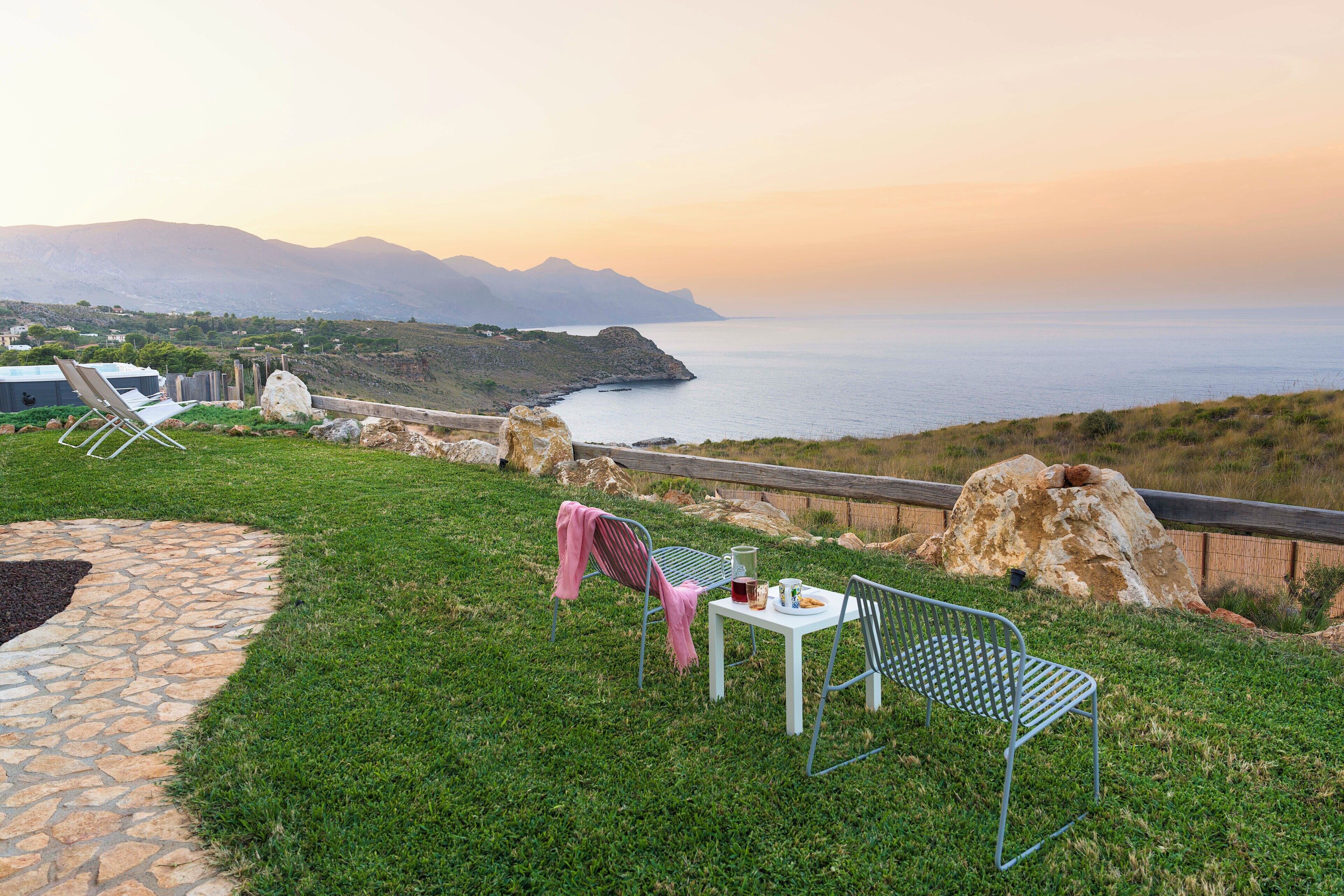 A scenic outdoor seating area with a table and chairs, overlooking a coastal landscape with mountains in the distance.