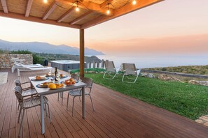 Une terrasse en bois avec une table dressée pour un repas, offrant une vue panoramique sur la mer et les montagnes.
