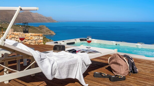 Een houten terras met een lounge stoel, een klein tafeltje met een wijnglas, een boek en een tas.
