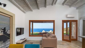 A living room with a large window offering a view of the sea, a sofa, a TV, and a dining area with chairs. - Villa Zingaro with sea view and pool (Castellammare del Golfo)