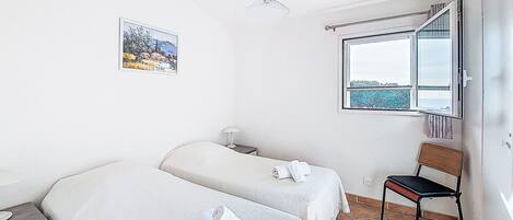 A bedroom with two beds, a window with a view, a chair, and a bedside table.