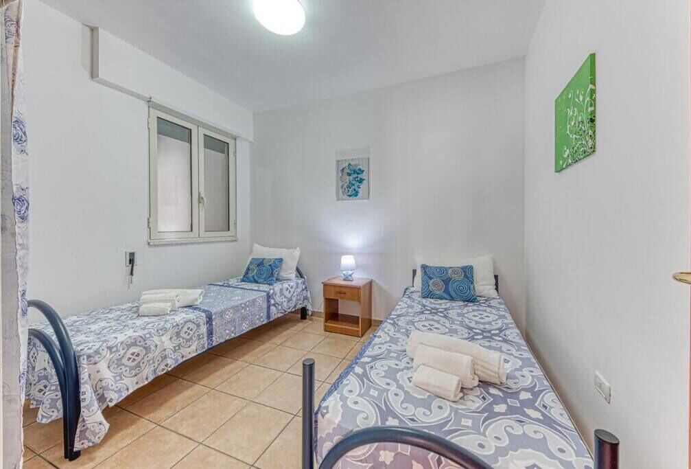 Quarto com duas camas de solteiro, janela, mesa de cabeceira e uma obra de arte na parede.