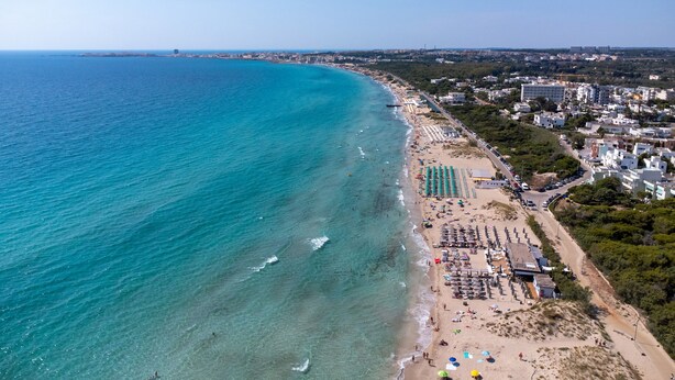 Luchtbeeld van een kuststrand met een rij strandgebouwen, een zandstrand met parasols en een heldere blauwe zee.