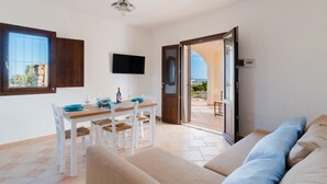 Een moderne woonkamer met een bank, een eettafel met stoelen, een aan de muur bevestigde televisie, en uitzicht op zee door een open deur.