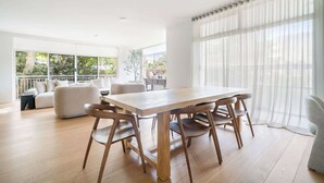 Een ruime woonkamer met een houten eettafel en stoelen, een bank en een groot raam met lichte gordijnen.