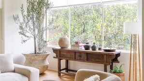 Een gezellige woonkamer met een groot raam, een houten bureau, een olijfboompje in pot en een moderne lamp.