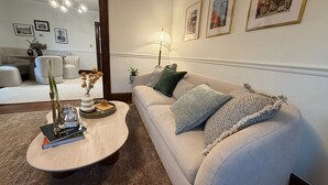 Un salon avec un canapé beige, une table basse et une lampe. Des tableaux encadrés ornent le mur, et un miroir reflète la pièce.
