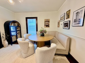 A dining area with a round table, four chairs, a bench, a bookshelf, and framed pictures on the wall.