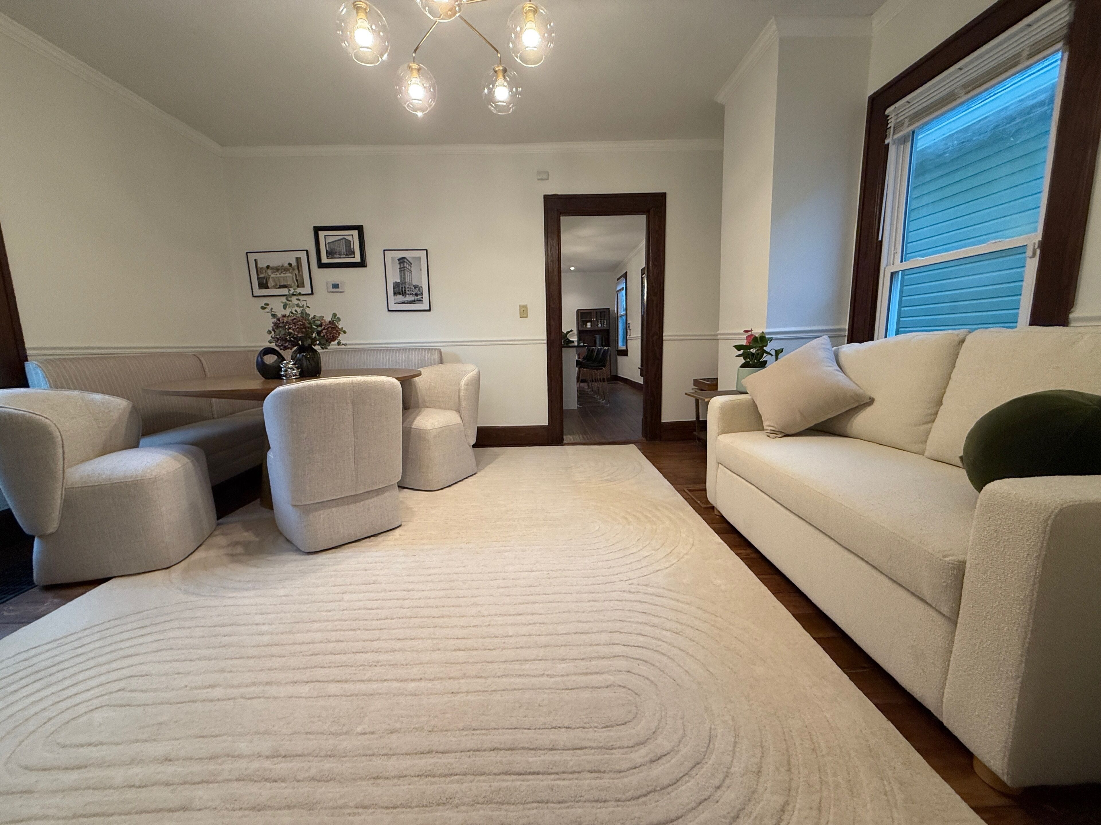 Sala de estar con un sofá color beige, dos sillones, una mesa de centro y un tapete estampado. Se observa una araña con varias lámparas y cuadros en la pared.