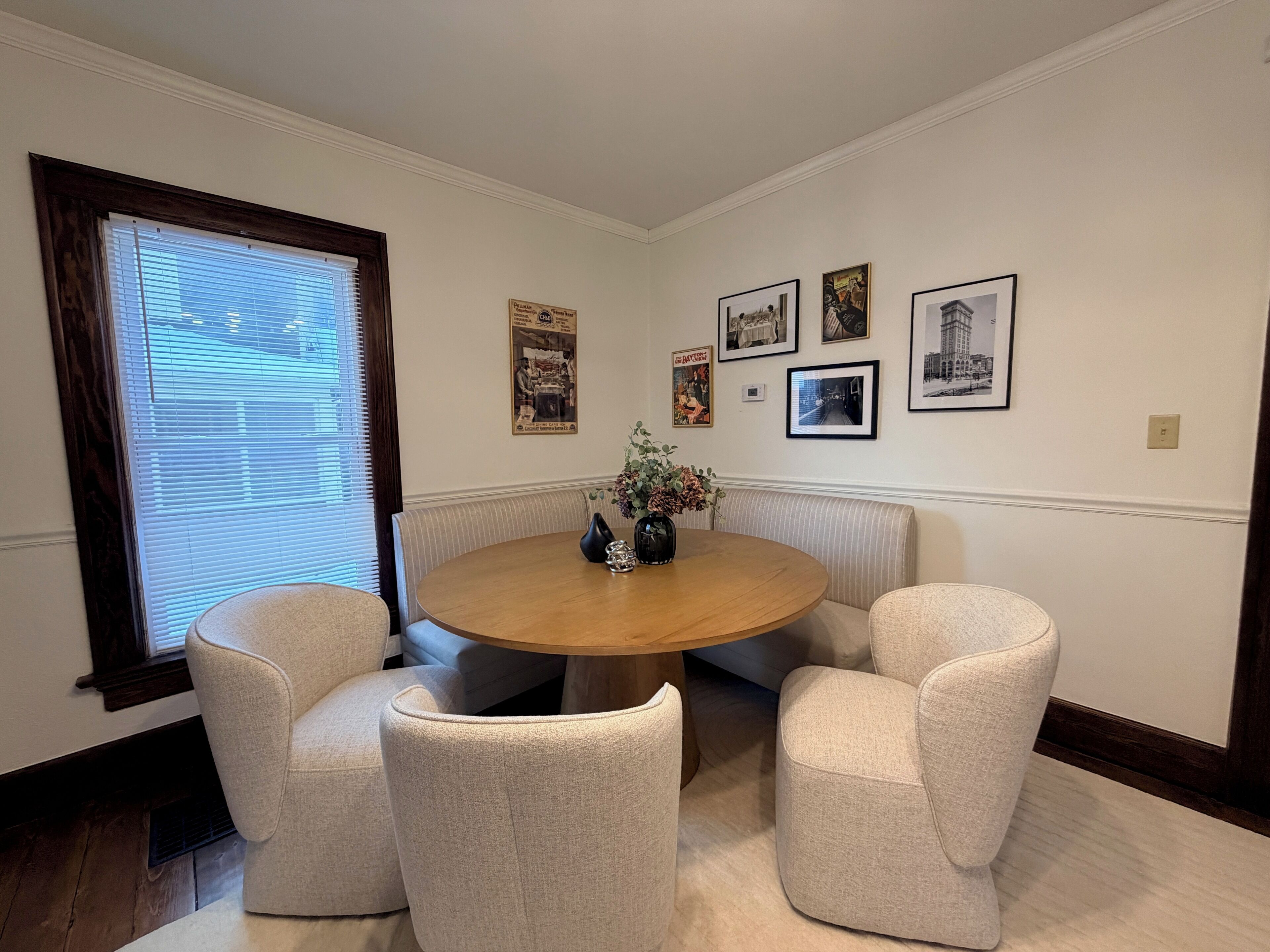 A dining area with a round table, four chairs, a vase with flowers, and framed pictures on the wall.
