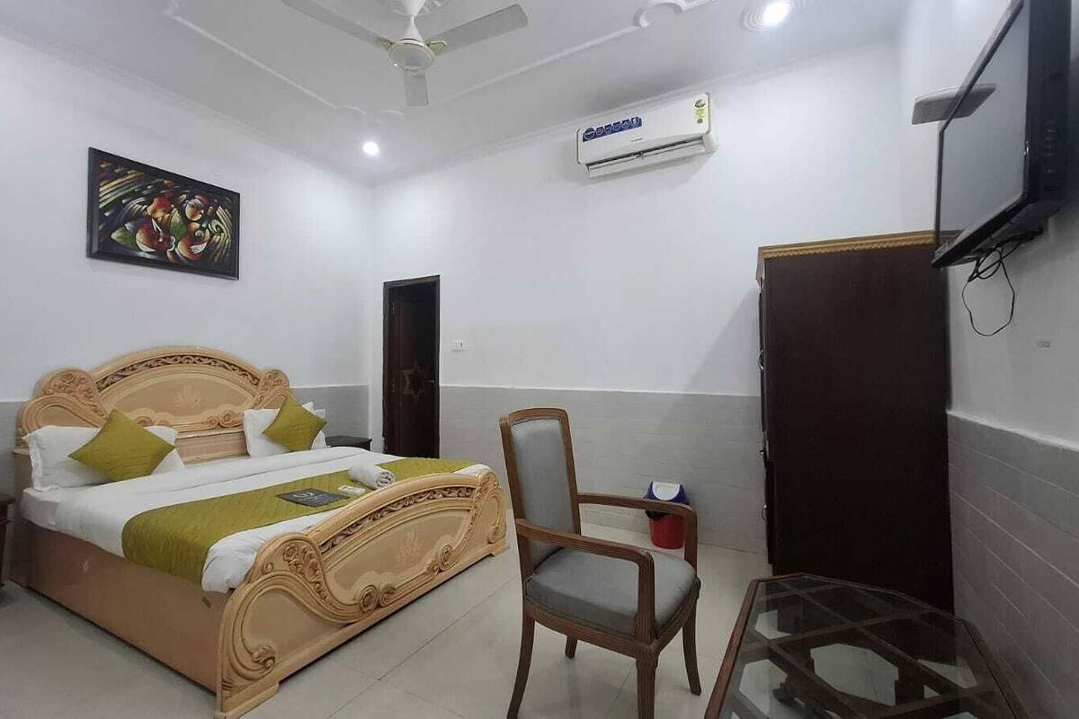 A bedroom with a bed, a chair, a desk, a TV, and an air conditioning unit.