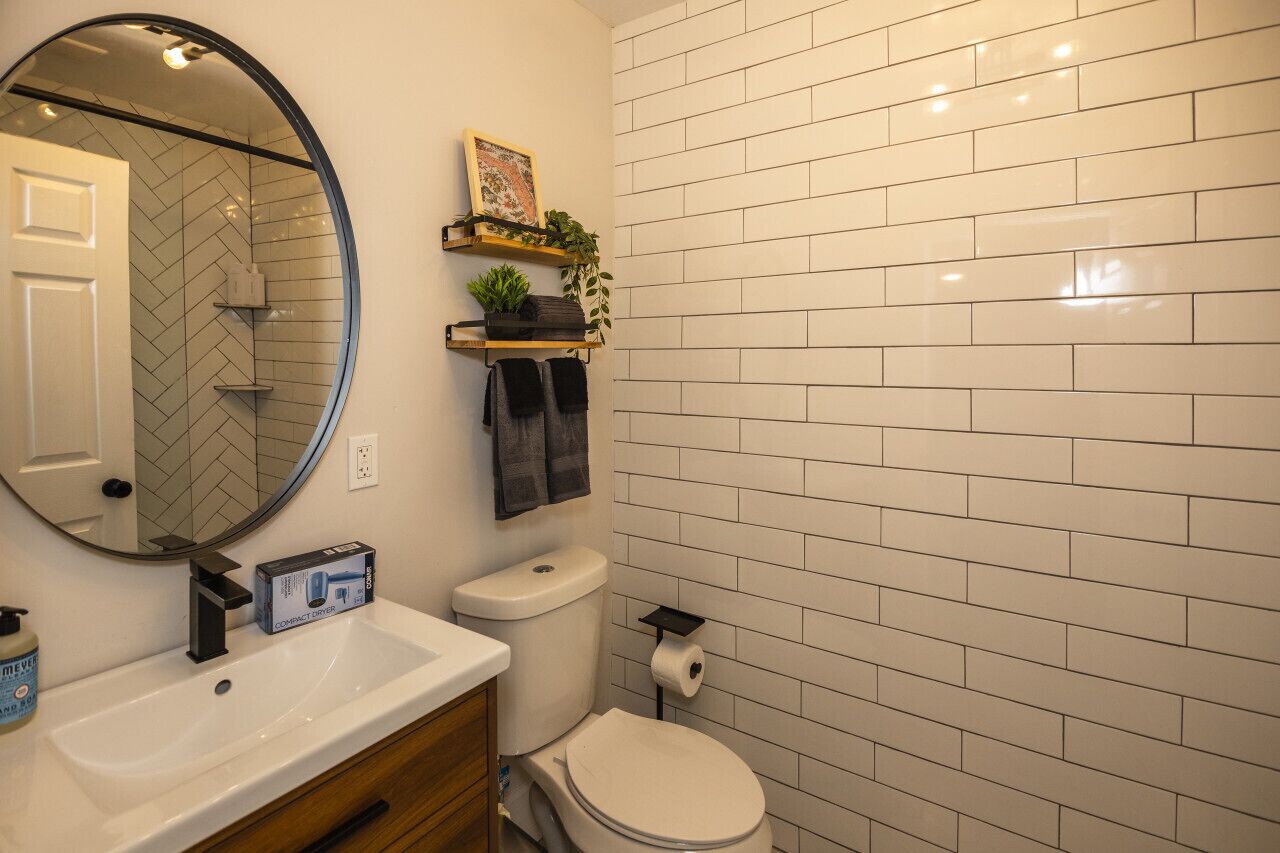 Ein Badezimmer mit weiß gefliestem Wandpaneel im Fischgrätmuster, einem runden Spiegel, einer weißen Toilette, einem weißen Waschbecken mit Seifenspender und einem Regal mit Pflanzen und Handtüchern.