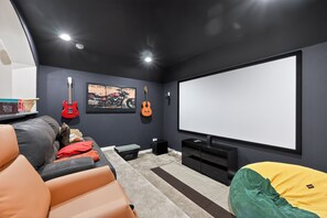 Una sala de cine en casa con una pantalla grande, dos guitarras, un sillón de peluche y un sofá.