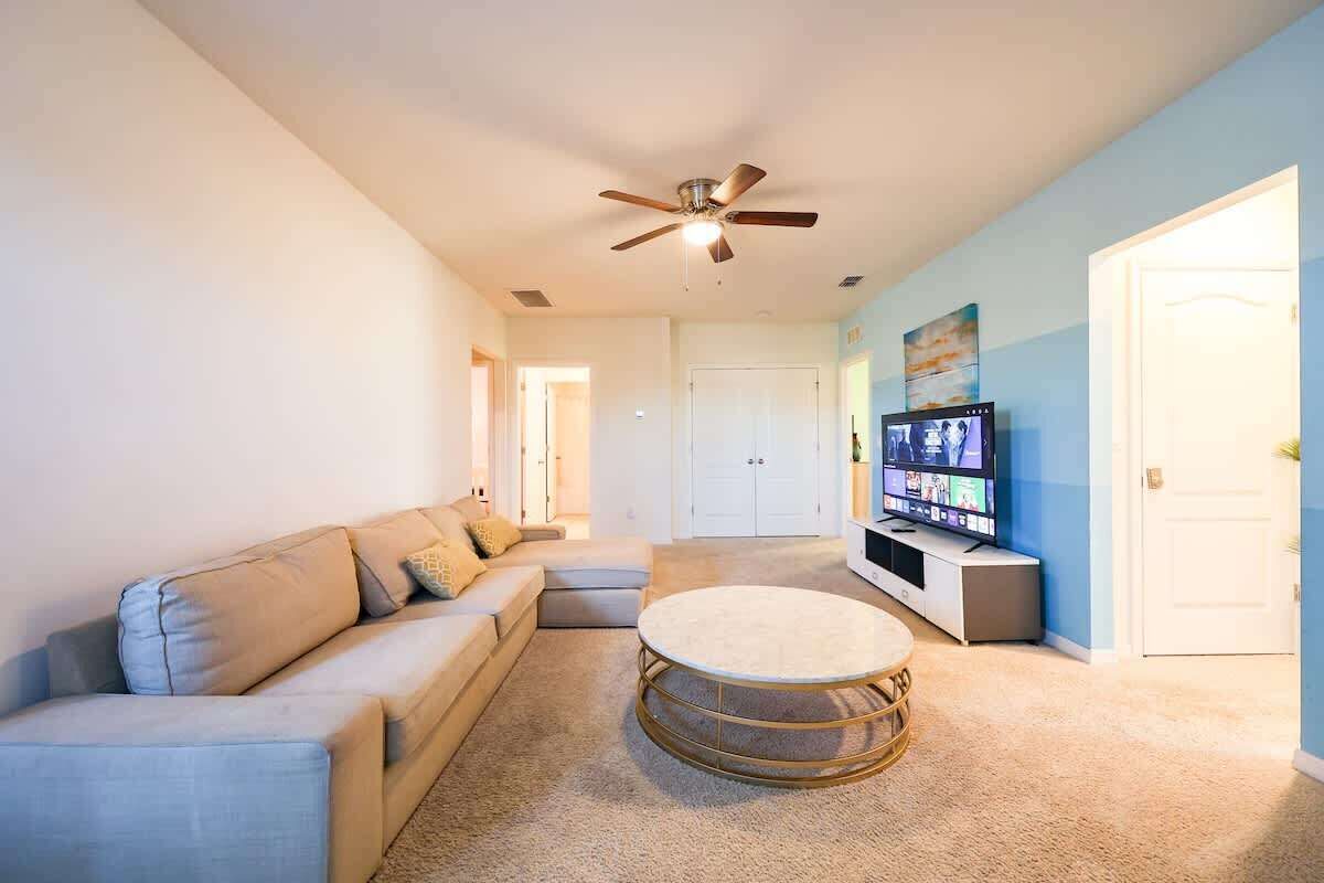 Un salon avec un canapé d’angle, une table basse ronde, un ventilateur de plafond, un téléviseur à écran plat et une porte visible au fond.