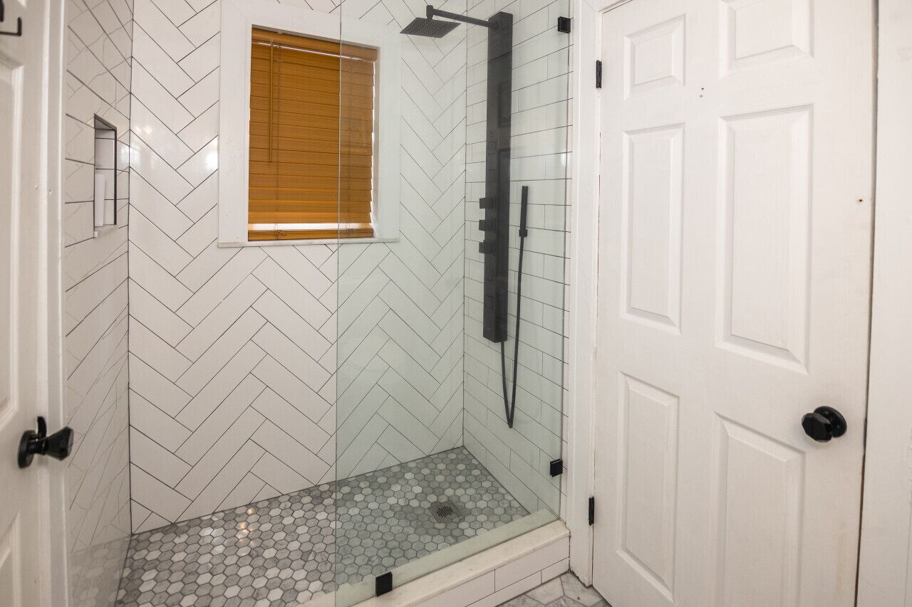 Un espace douche doté d’une porte en verre, de carreaux disposés en motif chevron sur les murs et d’une fenêtre avec des stores.