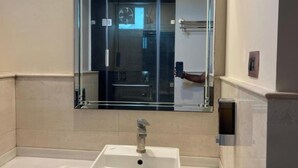 A bathroom with a white sink, a large mirror, and a glass door leading to a shower area. - Hotel Grand Tushar Elegant (Ghaziabad)