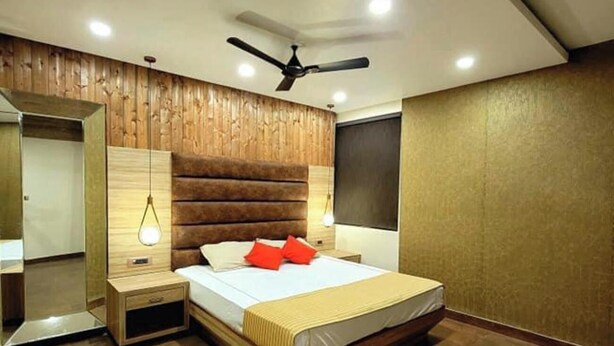 A modern bedroom with a wooden headboard, a bed with red pillows, a nightstand, a mirror, and a ceiling fan. - Hotel Grand Tushar Elegant (Ghaziabad)