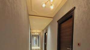 A hallway with a marble floor, a dark wooden door on the right, and a ceiling light fixture. - Hotel Grand Tushar Elegant (Ghaziabad)