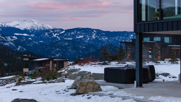 Een modern gebouw met grote ramen, een stenen terras en uitzicht op besneeuwde bergen.
