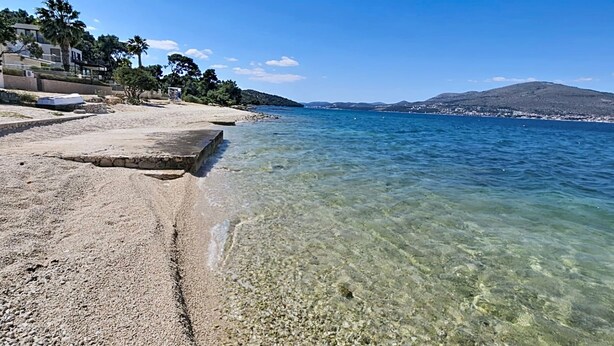 Playa de guijarros con agua cristalina, una plataforma de piedra y una cadena montañosa distante.