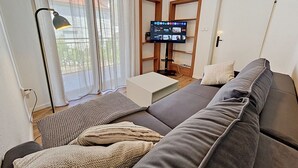 Una sala de estar con un sofá gris, un televisor de pantalla plana, una lámpara de pie y una ventana con cortinas translúcidas.