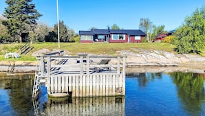 A wooden dock extending into a calm body of water with a clear sky. In the background, there is a red building with a dark roof and a flagpole with a flag. - Havstomten by Interhome (Blekinge)