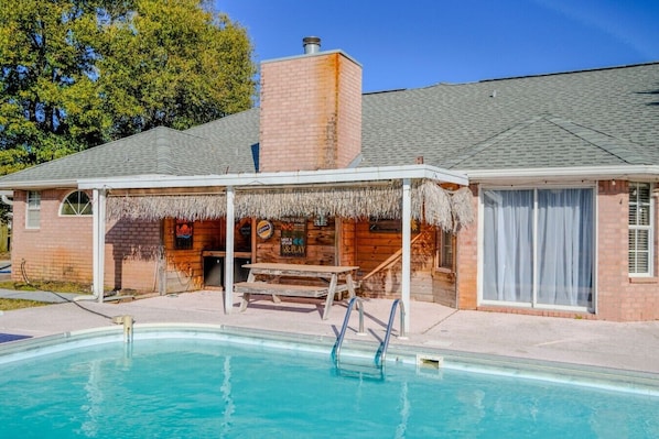 Un espace aménagé au bord de la piscine, avec un coin salon abrité, un bâtiment en briques et une piscine à l’eau d’un bleu limpide.
