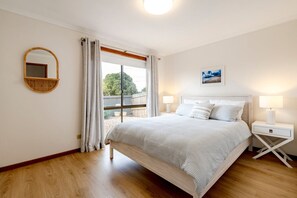 Ein Schlafzimmer mit einem großen Bett, einem Nachttisch mit Lampe, einem wandmontierten Spiegel und einem Fenster mit Vorhängen.