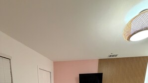 Une pièce avec un téléviseur fixé au mur, un meuble en bois, un pouf en osier et une grande fenêtre avec des stores.