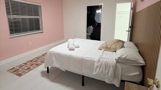 Une chambre à coucher comprenant un lit, une table de chevet, une fenêtre avec des stores, une porte et un petit tabouret en bois.