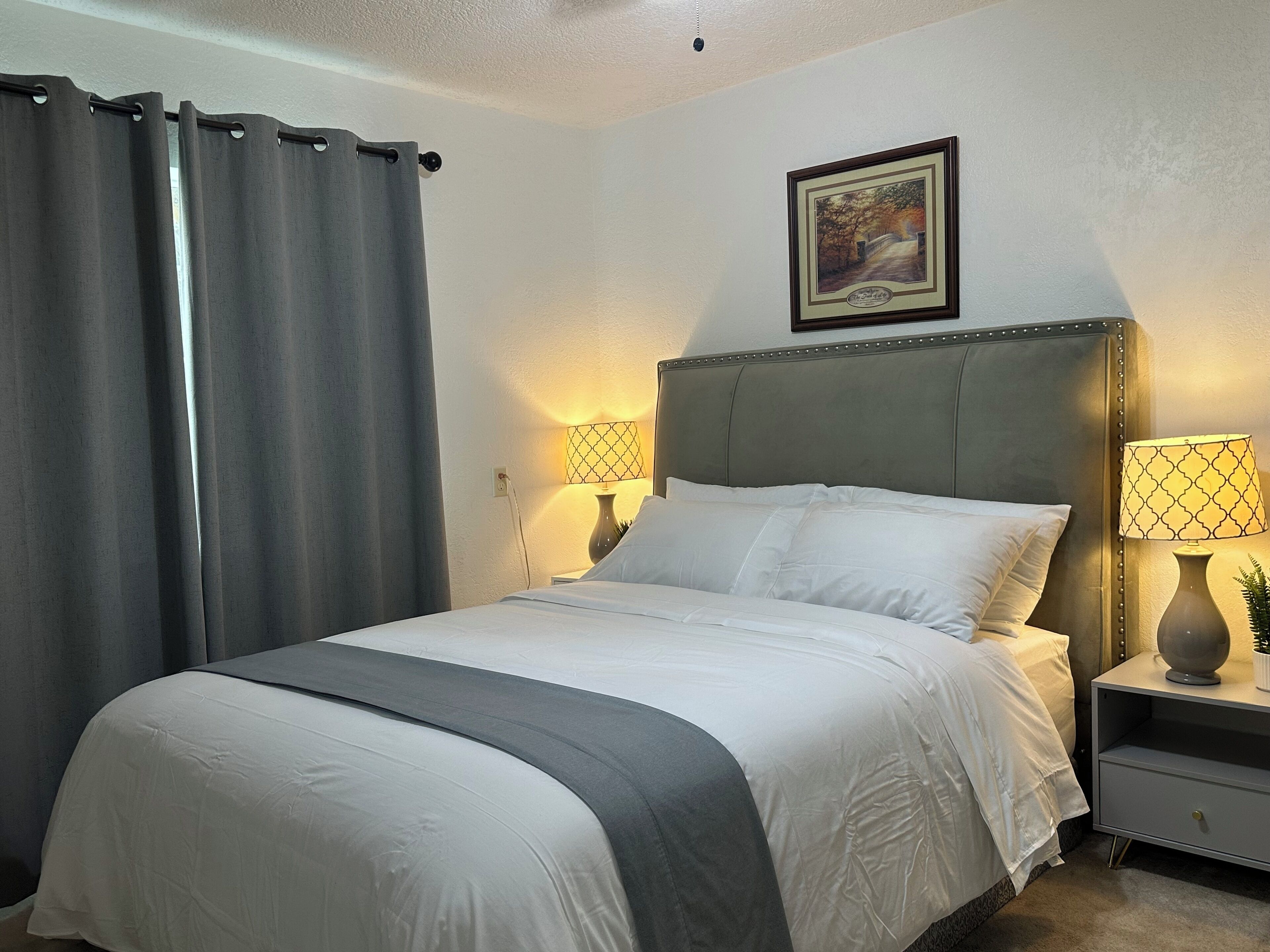 A neatly made bed with white linens and a grey accent, two bedside lamps, a framed picture on the wall, and a grey curtain.