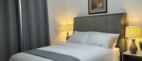 A neatly made bed with white linens and a grey accent, two bedside lamps, a framed picture on the wall, and a grey curtain.