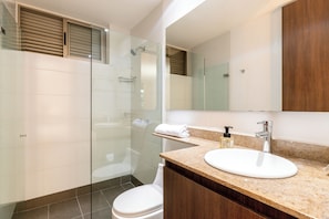 Un baño moderno con ducha envuelta en vidrio, inodoro blanco y encimera de mármol con lavamanos y espejo.