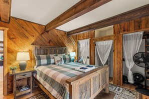 Une chambre avec un plafond en bois, un lit recouvert d’une couvre-lit à rayures, une table de chevet avec une lampe et une petite table de nuit avec des livres.